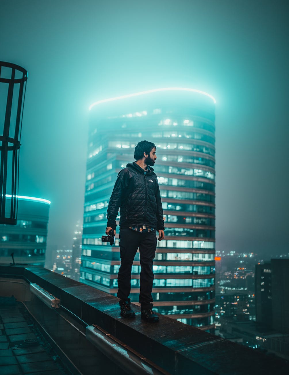 photo of man holding dslr camera walking on edge of building overlooking at night